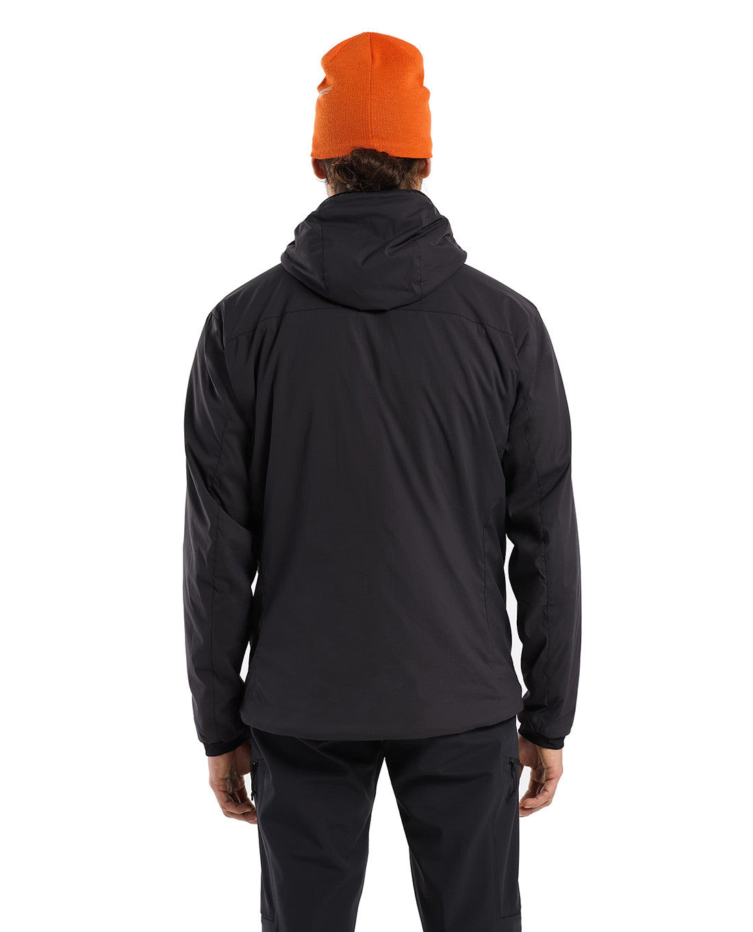 Eine Person steht mit dem Gesicht zur Wand und trägt ein ARC'TERYX PRO Arc'teryx Practitioner AR Hoody Men's in Carbon Copy, eine schwarze Hose und eine orangefarbene Mütze vor einem schlichten weißen Hintergrund.