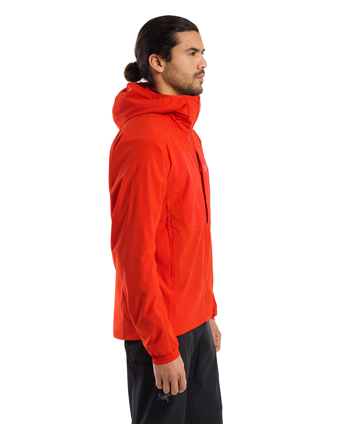 Ein Mann mit langen dunklen Haaren steht im Profil und trägt das ARC'TERYX PRO Arc'teryx Practitioner AR Hoody Men's - Dynasty in leuchtendem Rot mit schwarzer Hose vor einem schlichten weißen Hintergrund.