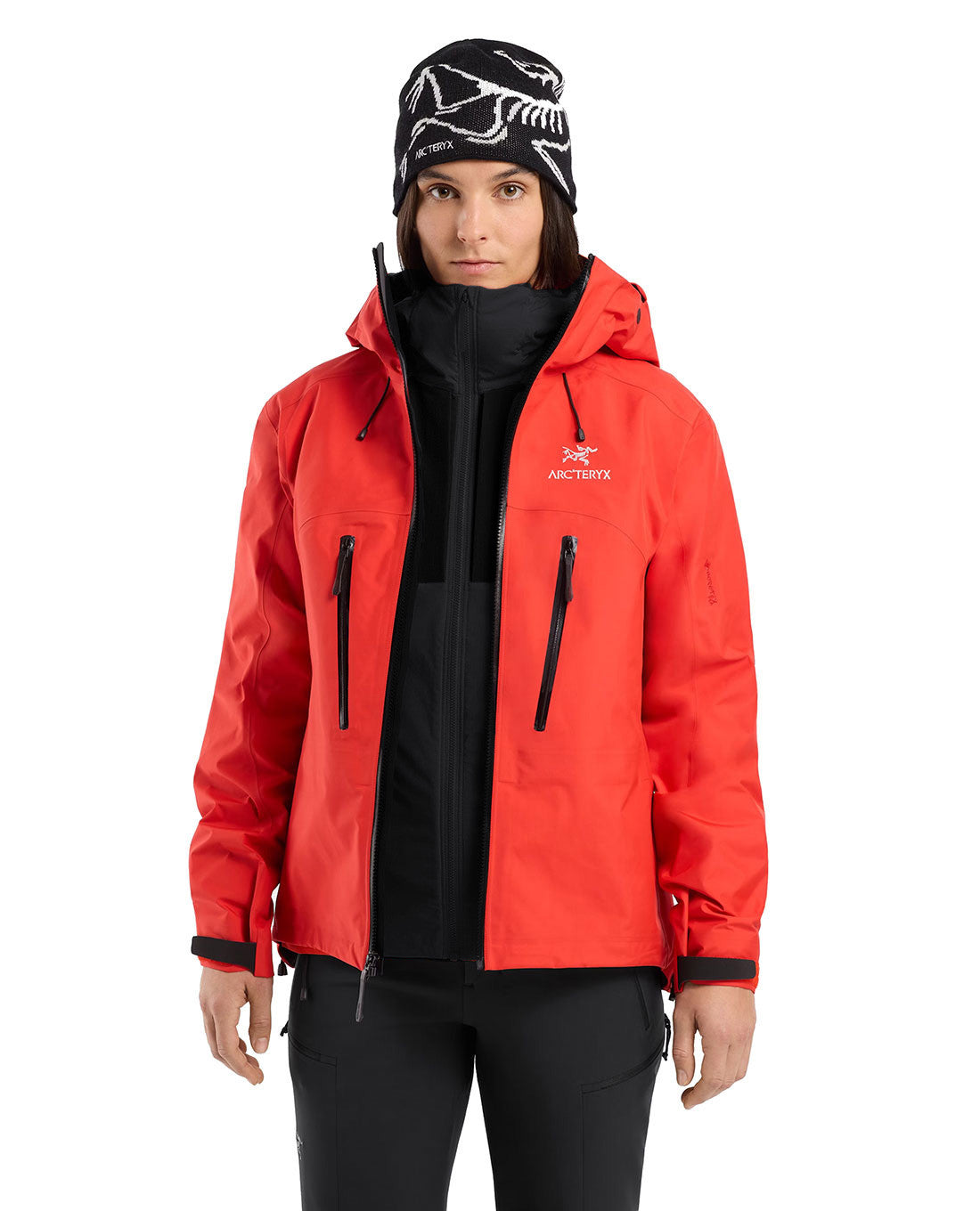 Eine Person im Arc'teryx Practitioner AR Hoody Women's - Carbon Copy von ARC'TERYX PRO, mit schwarzem Reißverschluss, schwarzer Hose und einer schwarz-weiß gemusterten Mütze, steht vor einem schlichten weißen Hintergrund.