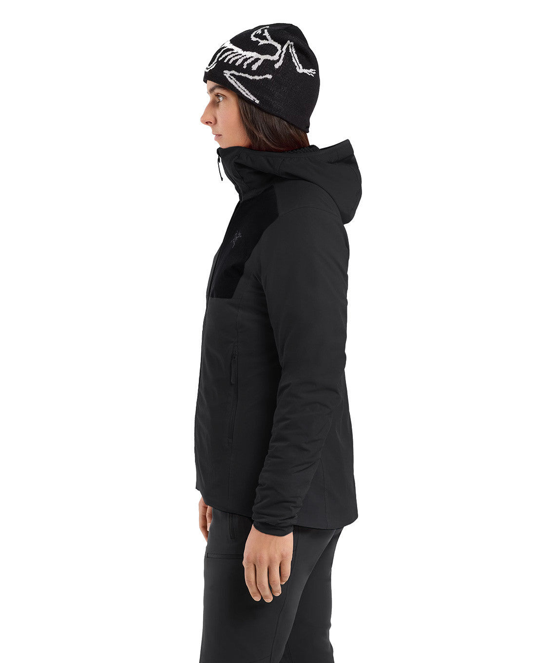 Eine Person in einem Arc'teryx Practitioner AR Hoody Women's - Carbon Copy by ARC'TERYX PRO und einer schwarzen Mütze mit weißem Skelettmuster steht im Profil vor einem schlichten weißen Hintergrund.