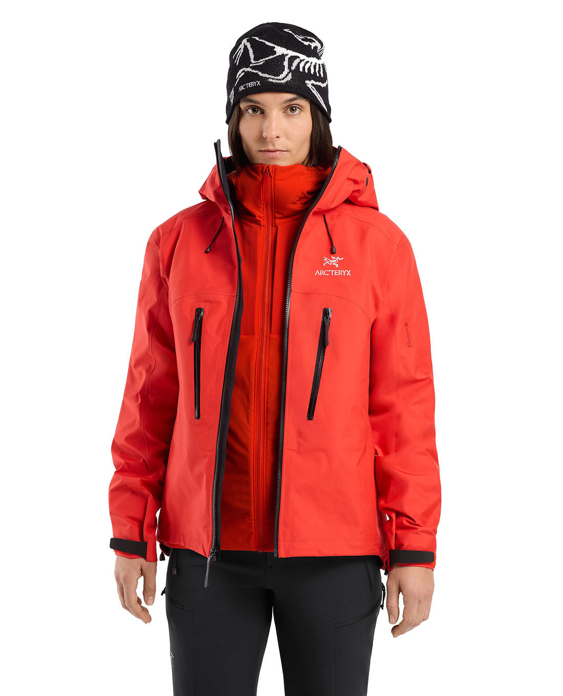 Eine Frau in einem roten ARC'TERYX PRO Practitioner AR Hoody Women's - Dynasty mit schwarzen Reißverschlüssen und einer schwarz-weißen Mütze steht mit neutralem Gesichtsausdruck vor einem weißen Hintergrund.