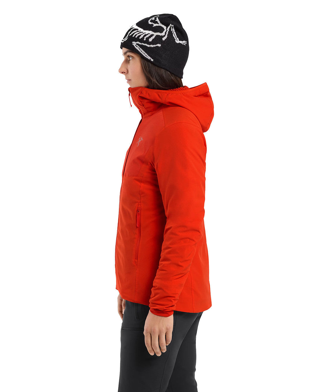 Eine Person im Profil trägt das leuchtend rote ARC'TERYX PRO Arc'teryx Practitioner AR Hoody Women's - Dynasty, eine schwarze Hose und eine schwarze Strickmütze mit weißem Skelettmuster vor einem schlichten weißen Hintergrund.
