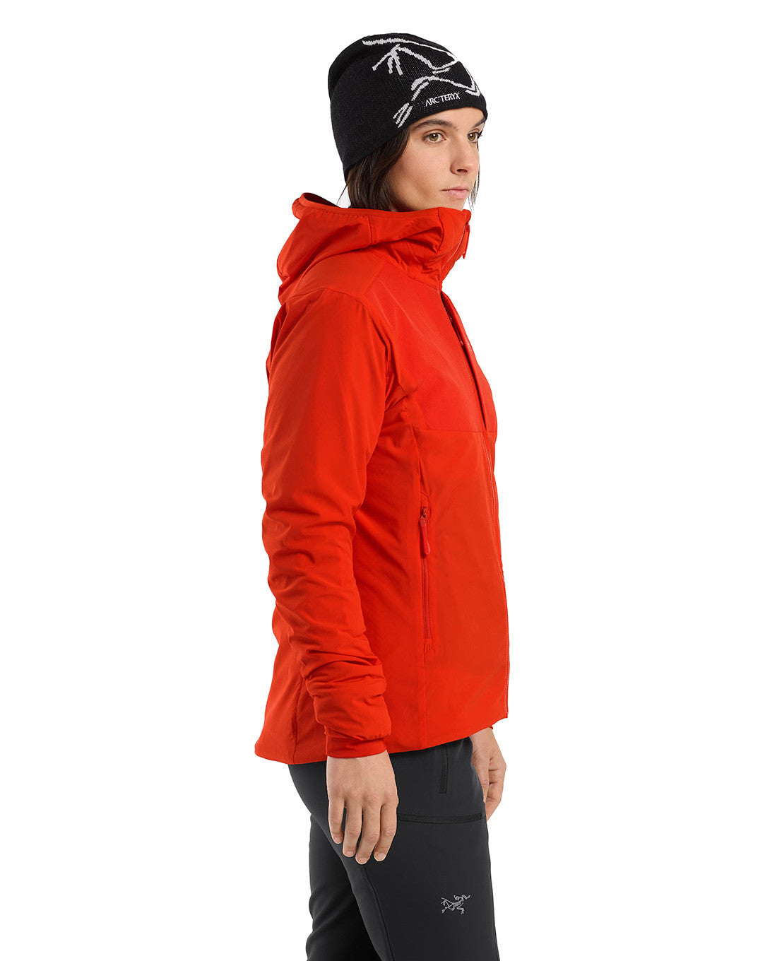 Eine Frau steht seitlich vor einem schlichten weißen Hintergrund und trägt ein ARC'TERYX PRO Arc'teryx Practitioner AR Hoody Women's - Dynasty in leuchtendem Rot, gepaart mit einer schwarzen Hose und einer schwarzen Mütze mit weißem Muster.
