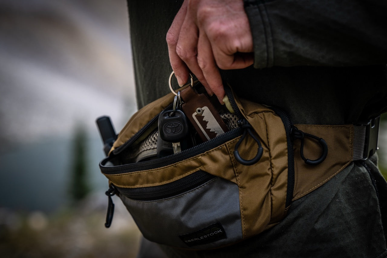 Eine Nahaufnahme zeigt eine Hand, die in die EBERLESTOCK Bando Bag Standard greift, um Schlüssel und ein kleines Multitool aus dem CCW-Fach zu holen, vor einem unscharfen Außenhintergrund.