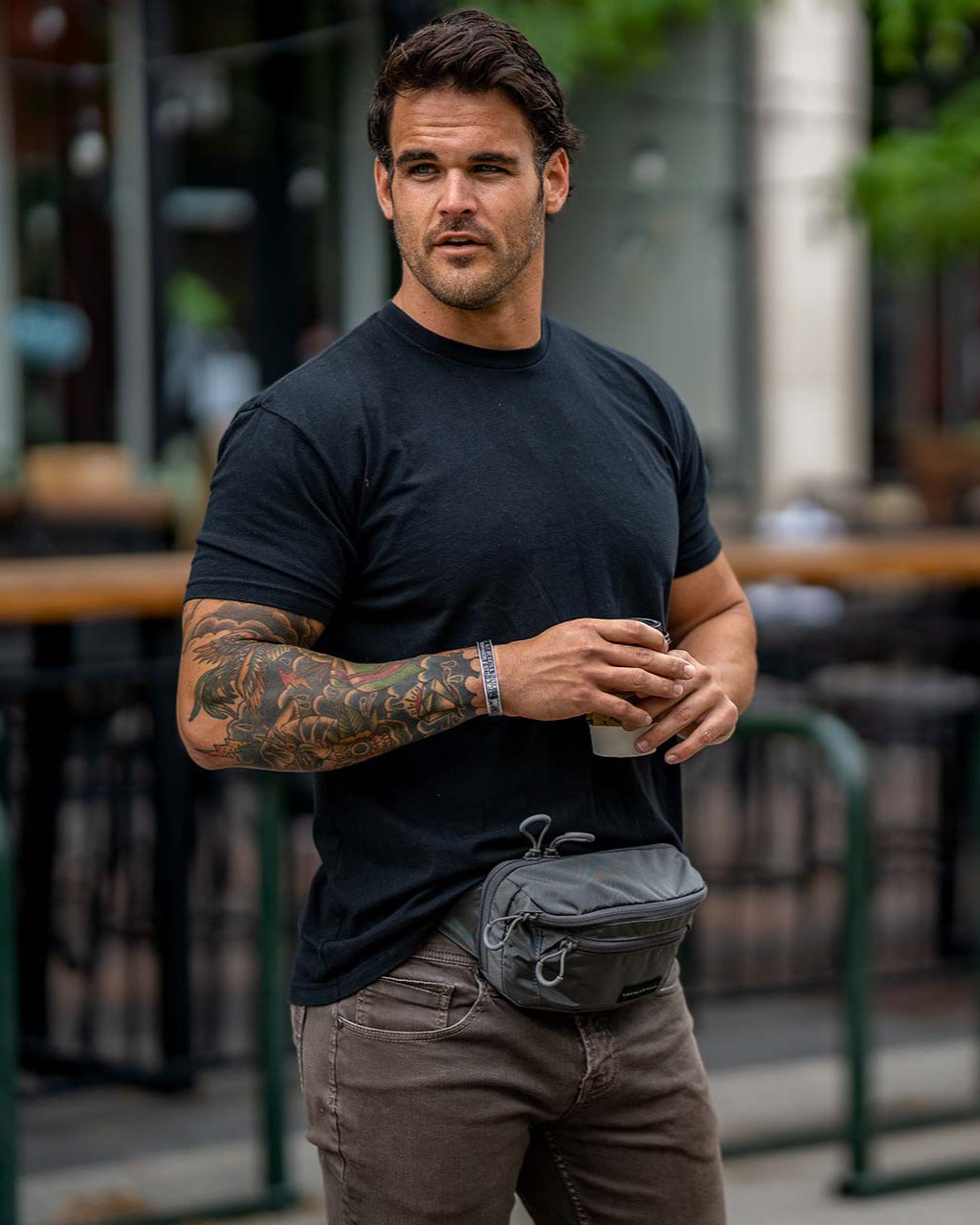 Ein bärtiger Mann mit dunklen Haaren und einer Tätowierung am rechten Arm steht im Freien, trägt ein schwarzes T-Shirt, eine braune Hose und eine EBERLESTOCK Bando Bag XL zum verdeckten Tragen. Im Hintergrund sind Bäume und ein Gebäude zu sehen.
