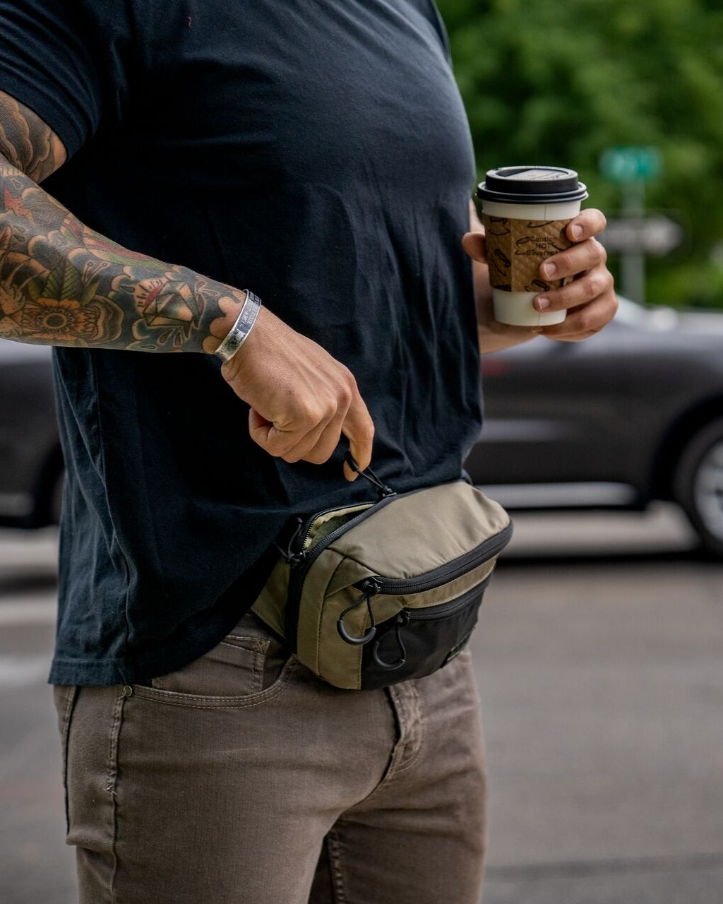 Eine tätowierte Person hält einen Kaffee in der Hand und öffnet den Reißverschluss der EBERLESTOCK Bando Bag XL, einer taktischen Umhängetasche, die sich ideal zum verdeckten Tragen eignet. Sie steht an einer Straße mit verschwommenen Autos und Grünflächen im Hintergrund.