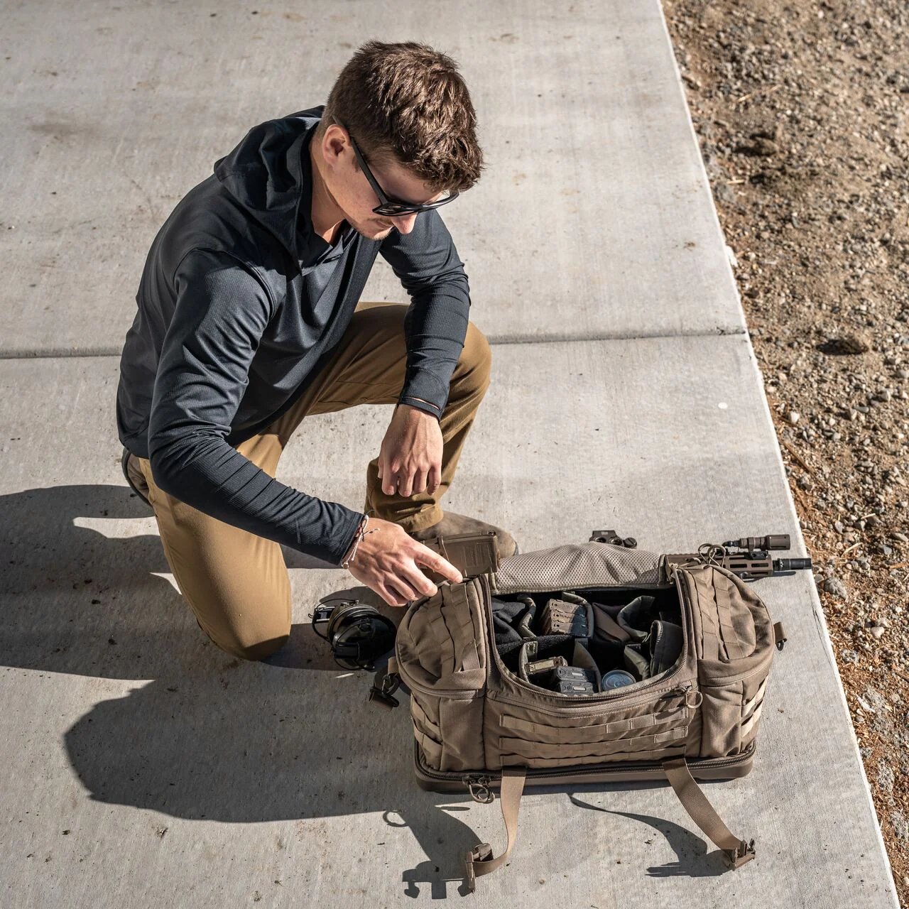 Ein Mann mit Sonnenbrille, dunklem Kapuzenpulli und hellbrauner Hose kniet auf dem Beton und sortiert Handschuhe und Ausrüstung in einer hellbraunen EBERLESTOCK Eberlestock Bang Bag Schießstandtasche, während das Sonnenlicht Schatten auf den Boden wirft.