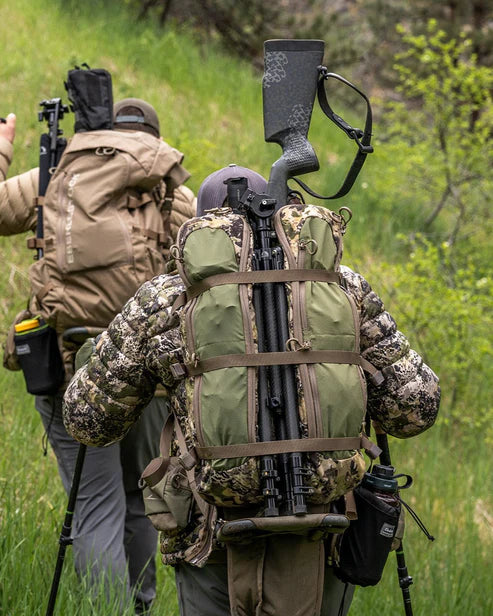 Zwei Wanderer in Tarnjacken, jeder mit einem großen Rucksack - darunter ein EBERLESTOCK Batwing Large - Dry Earth - wandern durch grasbewachsenes Terrain. Ein Rucksack trägt ein Gewehr, beide Wanderer benutzen Trekkingstöcke.