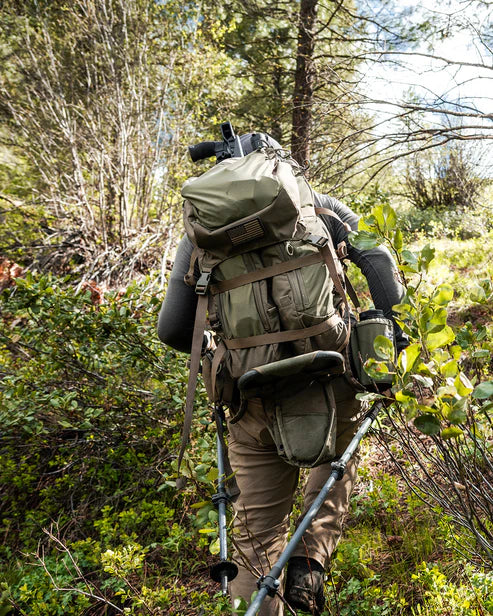 Mit einem EBERLESTOCK Eberlestock Batwing Large - Military Green und Trekkingstöcken wandert ein Wanderer unter dem sonnigen Himmel bergauf durch den dichten grünen Wald und schlängelt sich zwischen Bäumen und Sträuchern hindurch.