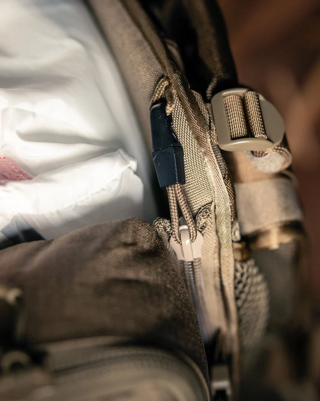 Nahaufnahme einer EBERLESTOCK Eberlestock Batwing V2 in Braun, die den Reißverschluss, die Schnalle und einen weißen Gegenstand zeigt, der teilweise in der Außentasche verstaut ist. Die Aufnahme hebt den strukturierten Stoff und die detaillierten Verschlüsse der Tasche hervor.