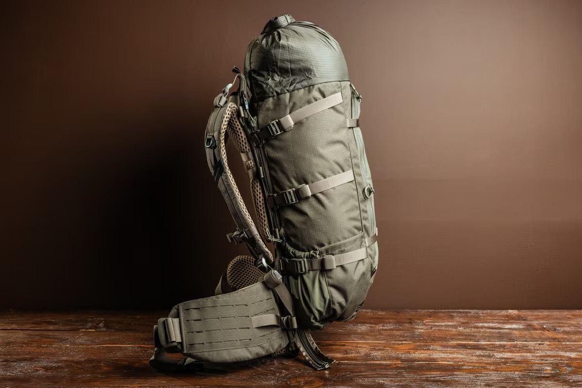 Ein EBERLESTOCK Brooks 3500 Wanderrucksack in Military Green mit mehreren Gurten und gepolsterten Schulter-/Taillenstützen steht aufrecht auf einer Holzfläche vor einem schlichten braunen Hintergrund.
