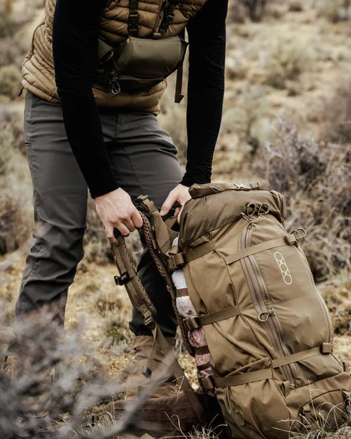 Eine Person in Outdoor-Ausrüstung stellt einen EBERLESTOCK Brooks 3500 - Military Green Rucksack auf dem Boden in einer trockenen, grasbewachsenen Landschaft mit Büschen ein. Ihr Kopf befindet sich außerhalb des Rahmens.