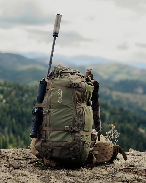 Ein EBERLESTOCK Brooks 3500 - Military Green Rucksack mit einem Gewehr mit Zielfernrohr sitzt auf einem Felsvorsprung und überblickt eine weite, bewaldete Berglandschaft unter einem wolkenverhangenen Himmel.