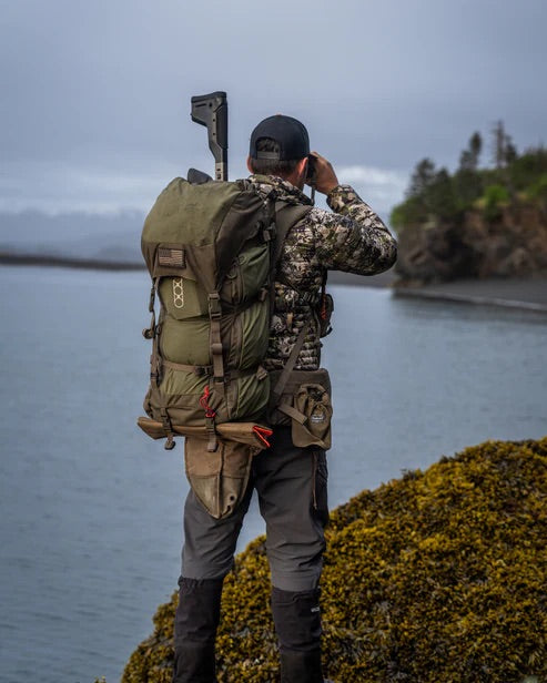 In Outdoor-Ausrüstung und mit dem EBERLESTOCK Brooks 3500 Rucksack in Military Green steht eine Person auf einem moosbewachsenen Felsen am Wasser und beobachtet mit einem Fernglas die entfernten Bäume und Berge.