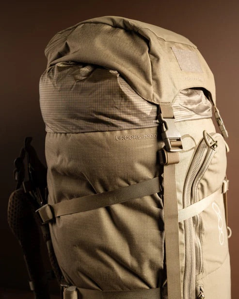 Eine Nahaufnahme eines EBERLESTOCK Brooks 7000 Rucksacks in Military Green mit mehreren Riemen und Reißverschlüssen vor einem schlichten braunen Hintergrund. Die geräumige Tasche ist ideal für Trekking- und Jagdabenteuer.