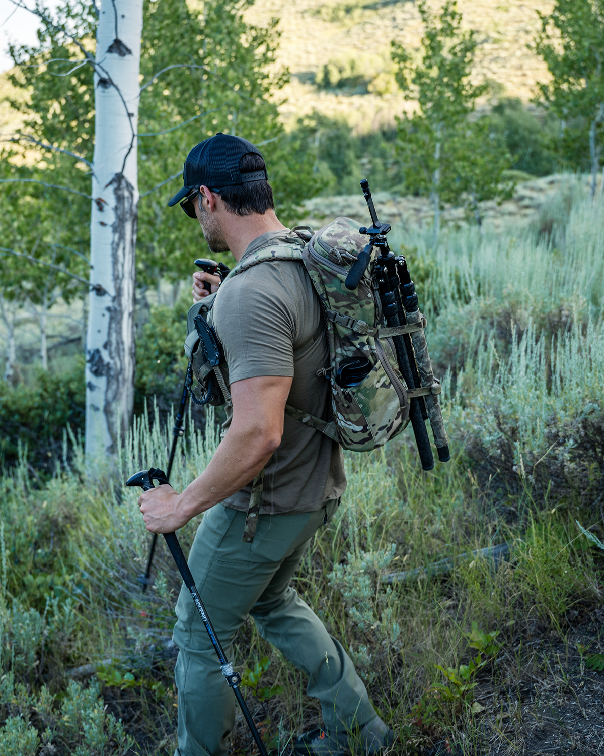 Ein Wanderer mit Mütze und Sonnenbrille wandert mit Trekkingstöcken und einem EBERLESTOCK Brute Scout Tagesrucksack in Camouflage mit montiertem Stativ durch ein grasbewachsenes Waldgebiet. Bäume und grüne Vegetation vervollständigen die Outdoor-Szene.