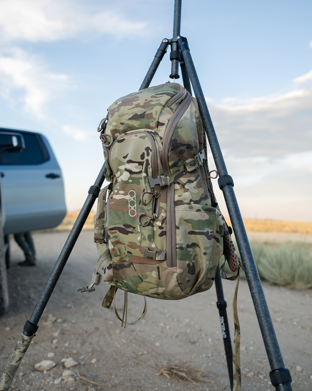 Der EBERLESTOCK Eberlestock Brute Scout Tagesrucksack im Camouflage-Stil hängt an einem schwarzen Stativ auf einer Schotterstraße, mit geparktem Auto und grüner Landschaft im Hintergrund unter teils bewölktem Himmel.