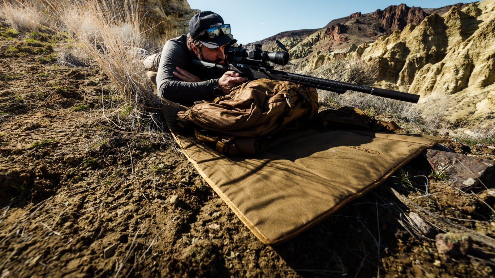 Bekleidet mit Outdoor-Kleidung liegt jemand in Bauchlage auf der EBERLESTOCK Dirt Nap Shooting Mat in einer felsigen, trockenen Gegend und zielt mit einem Zielfernrohr, das auf einem braunen Rucksack ruht. Trockenes Gras und zerklüftete Hügel machen es ideal für Zielübungen.