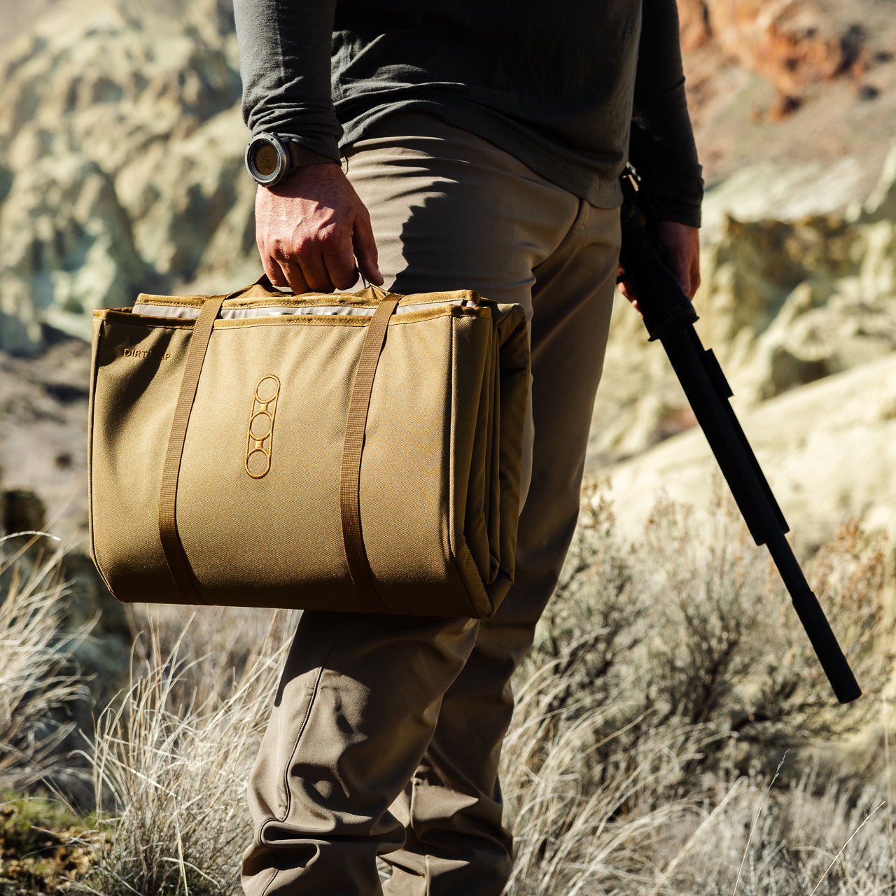 Eine Person in Outdoor-Ausrüstung steht auf felsigem Gelände mit schroffen Hügeln im Rücken und hält eine EBERLESTOCK Dirt Nap Shooting Mat in der Hand - ideal für das Schießtraining in der Wildnis.