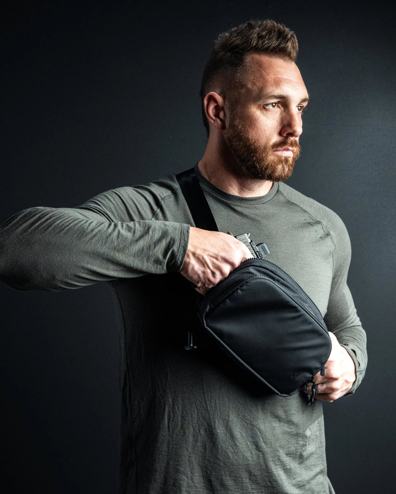 Ein bärtiger Mann in einem langärmeligen olivfarbenen Hemd zieht vor einem dunklen Hintergrund einen Gegenstand aus der EBERLESTOCK Fade Flex Crossbody Bag.