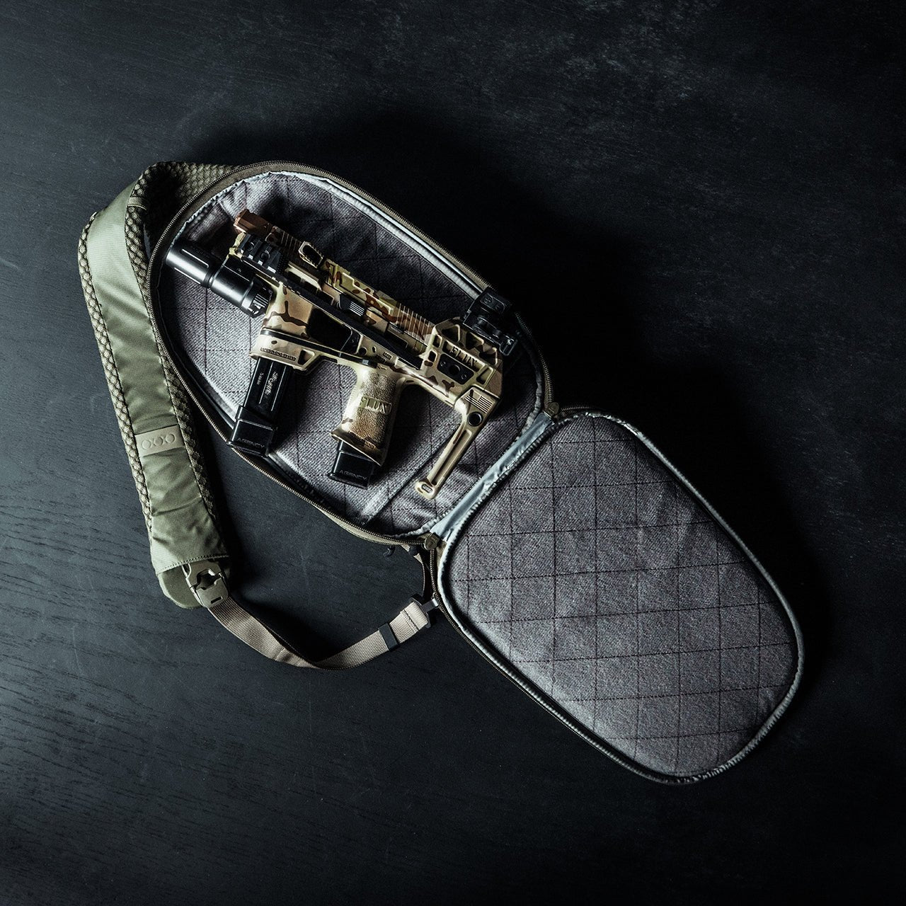 Eine kompakte, braun-schwarze Personal Defense Weapon mit zwei Magazinen befindet sich in einer grauen, gepolsterten EBERLESTOCK Fade Sling Tragetasche mit Riemen und Reißverschluss, die auf einer dunkel strukturierten Oberfläche präsentiert wird.