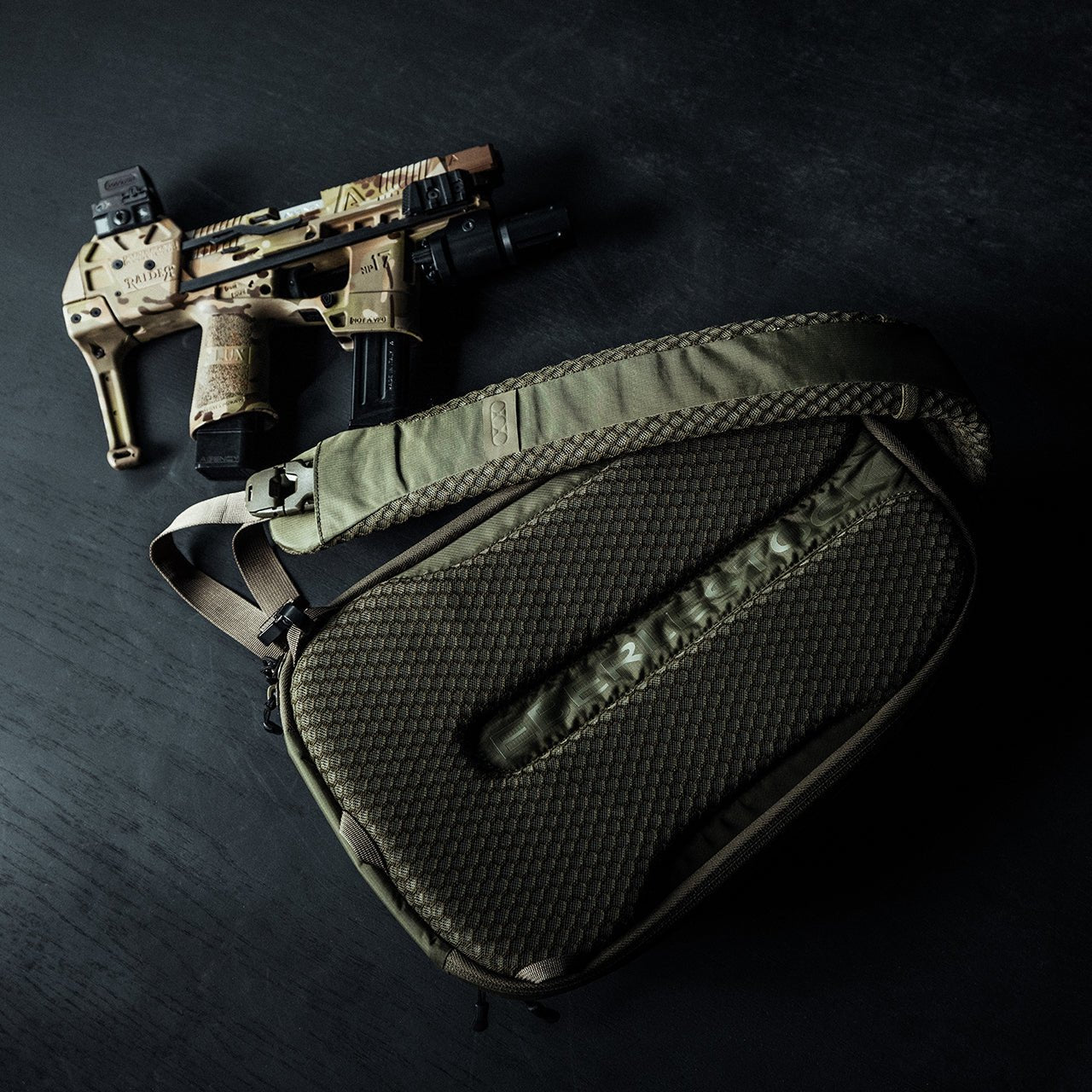 Eine hellbraune und schwarze taktische Pistole mit Aufsätzen liegt neben einem olivgrünen EBERLESTOCK Fade Sling, beide bereit für jede Situation auf einer dunklen Oberfläche - perfekt für Liebhaber von Waffen zur persönlichen Verteidigung.