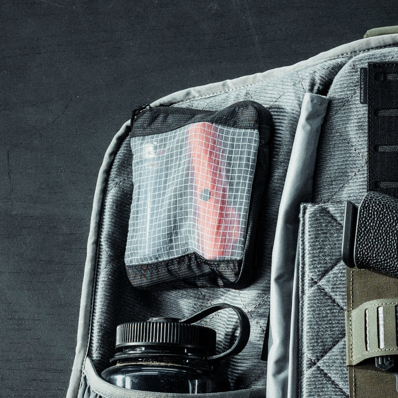 Die Nahaufnahme eines grauen Rucksacks zeigt eine EBERLESTOCK Fade View Pouch Small aus durchsichtigem Ripstop-Nylon, die einen roten Gegenstand hält, daneben eine schwarze Wasserflasche mit Schlaufendeckel sowie sichtbare Fächer und Gurte vor einem dunklen Hintergrund.
