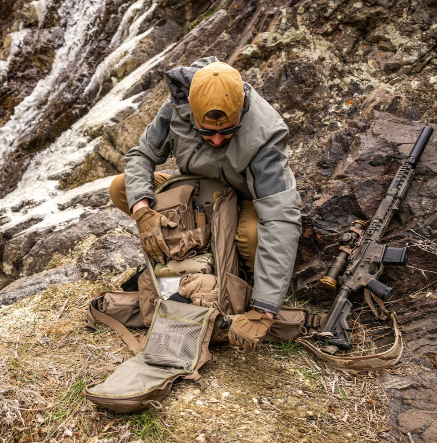 Bekleidet mit Outdoor-Ausrüstung und Handschuhen kniet eine Person auf einem felsigen Hügel und öffnet einen EBERLESTOCK FreeFall 2000 Pack. In der Nähe ruht ein Gewehr mit Zielfernrohr auf dem schneebedeckten Boden - eine Ausrüstung, die für Abenteuer in unwegsamem Gelände bereit ist.