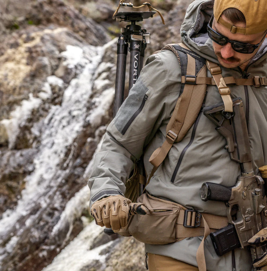 Eine Person in Outdoor-Ausrüstung, mit Handschuhen und Sonnenbrille passt einen EBERLESTOCK FreeFall 2000 Pack an, während sie ein Gewehr trägt; ein Stativ und ein Wasserfall unterstreichen die raue Landschaft in dieser Outdoor-Abenteuerszene.