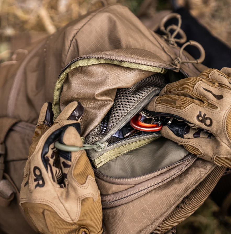 Handschuhe öffnen den braunen EBERLESTOCK FreeFall 2000 Pack und geben den Blick frei auf saubere Netztaschen und einen roten Karabiner im Inneren - ein Beweis für die outdoor-taugliche Organisation dieses vielseitigen Rucksacks.