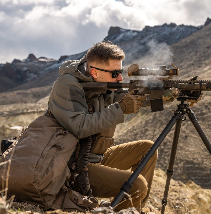 Eine Person mit Sonnenbrille und Outdoor-Ausrüstung richtet ein Gewehr auf einem Stativ in den Bergen, Rauch steigt aus dem Lauf auf. Neben ihnen steht der EBERLESTOCK FreeFall 2000 Pack - perfekt für jedes Outdoor-Abenteuer.