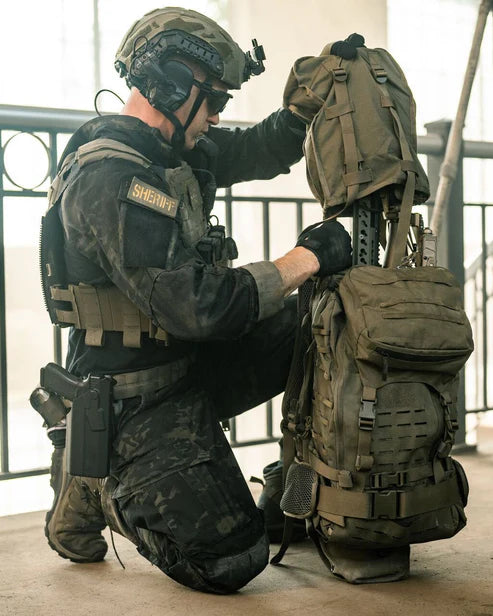 Ein Sheriff in taktischer Ausrüstung kniet in einem Haus, trägt einen Helm und eine Sonnenbrille und passt einen EBERLESTOCK Gunslinger II - Black Rucksack an. Die Industriekulisse besteht aus Metallgeländern und hellen Fenstern im Hintergrund.