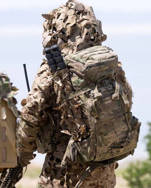 Eine Person in Camouflage und einem schwarzen EBERLESTOCK Eberlestock Gunslinger II Rucksack, von hinten gesehen, steht mit einem Funkgerät oder einer Antenne im Freien und fügt sich perfekt in die natürliche Umgebung ein.