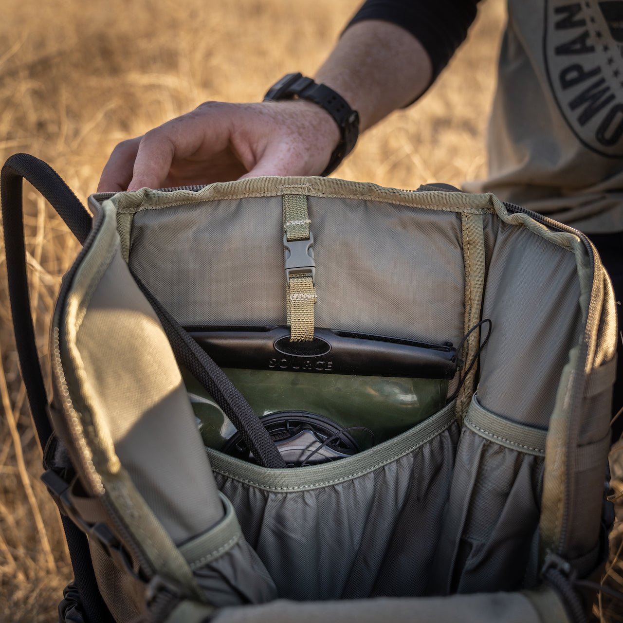 Im Freien öffnet jemand einen braunen Rucksack und bringt eine EBERLESTOCK Hydration System Bladder 2L und einen schwarzen Gegenstand im Innenfach zum Vorschein, während im Hintergrund trockenes Gras zu sehen ist.