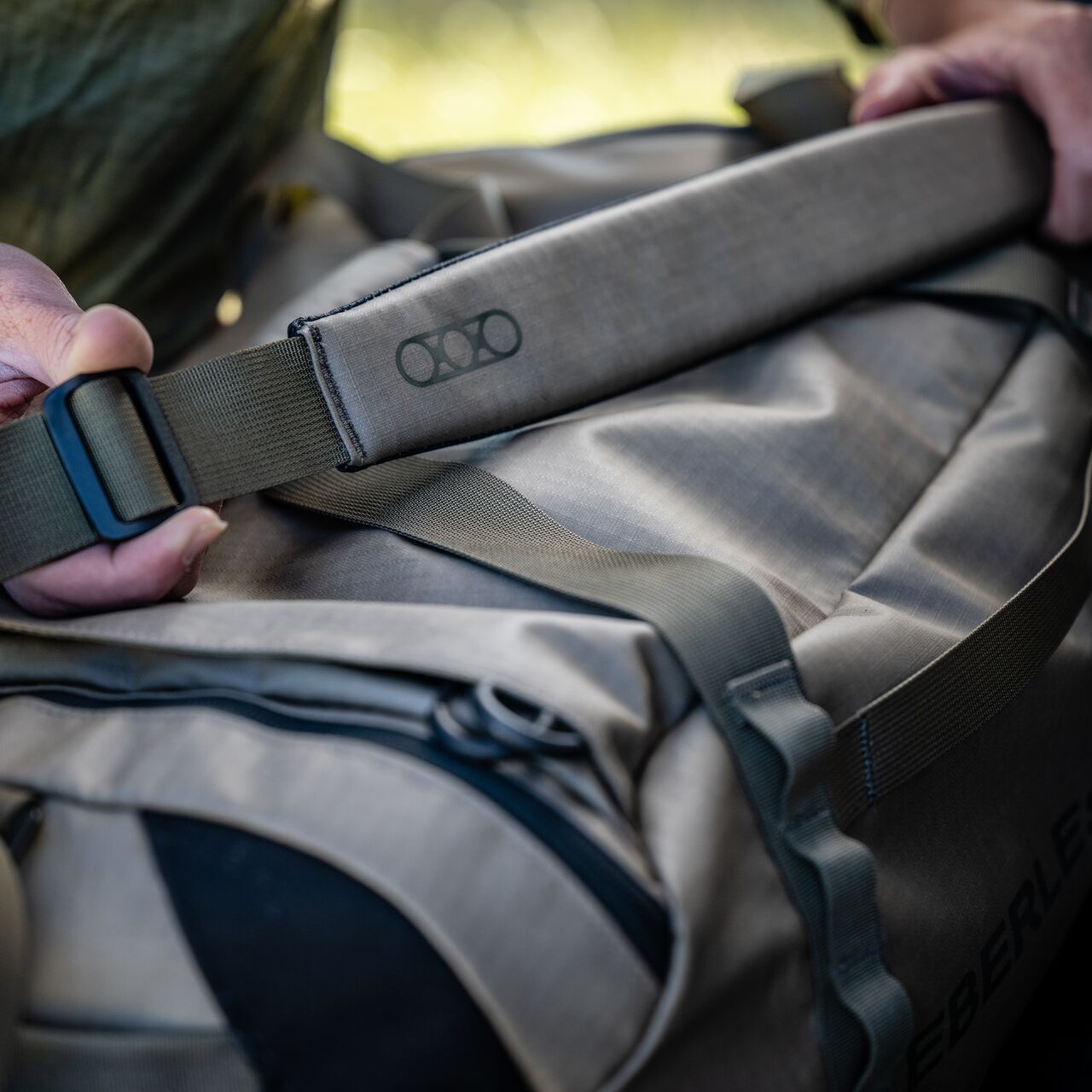 Nahaufnahme der Hände, die den Gurt einer EBERLESTOCK Hyllus Duffel 65 im Freien halten. Zu sehen sind der gepolsterte Schultergurt, der robuste Reißverschluss und das wasserfeste Material.