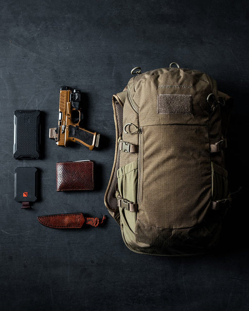 Ein EBERLESTOCK Eberlestock Jacknife - Dry Earth Rucksack liegt auf dunklem Untergrund neben einer Pistole, einem Messer mit braunem Griff, einem roten Wallet und zwei kompakten schwarzen Geräten.