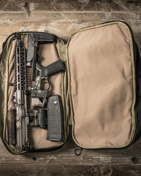 Ein zerlegtes schwarzes Gewehr mit einem Magazin liegt ordentlich in einem offenen, gepolsterten EBERLESTOCK Jacknife - Military Green Rucksack, einem 26-Liter-Koffer, der auf einer rustikalen Holzoberfläche ruht.