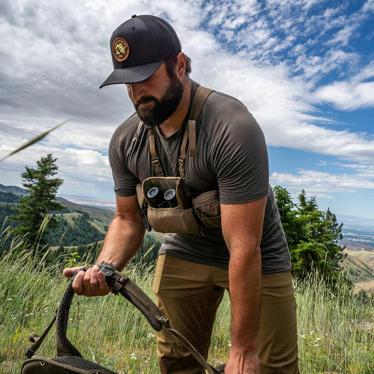 Ein bärtiger Mann mit Mütze und Outdoor-Ausrüstung steht in einer grasbewachsenen, bergigen Landschaft, trägt das EBERLESTOCK Lochsa Merino Short Sleeve - Black und hält seinen Rucksackgurt unter blauem Himmel und Wolken.