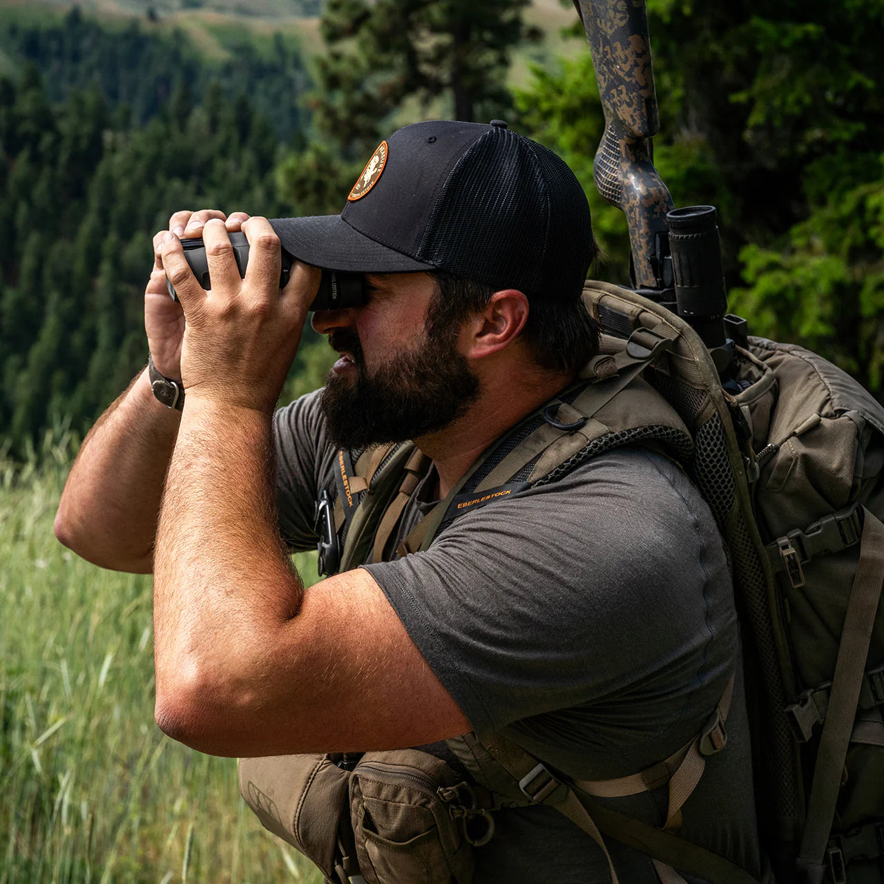Ein bärtiger Mann mit Mütze und EBERLESTOCK Lochsa Merino Short Sleeve - Loden sucht mit einem Fernglas einen grasbewachsenen Wald ab, ein Gewehr ist auf seinem Rucksack befestigt.