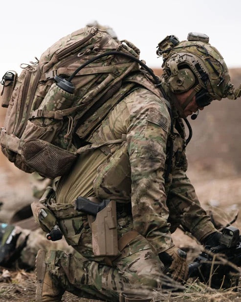Ein Soldat in Camouflage und mit Helm kniet im Freien und trägt einen EBERLESTOCK Lodrag II V2 - Dry Earth Rucksack und Ausrüstung, während er ein Gerät in unwegsamem Gelände bedient.