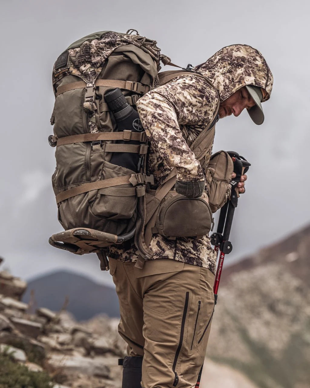 Eine Person in Camo-Outdoor-Ausrüstung und mit Mütze steht auf einem felsigen Pfad, trägt den EBERLESTOCK Mainframe - Tall Rucksack und hält Trekkingstöcke in der Hand, mit Bergen im Hintergrund.