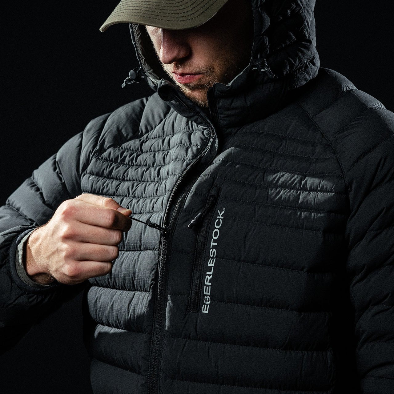 Eine Person in einem schwarzen Eberlestock McCall Down Hoody mit Kapuze und Mütze, die vorne mit einem Reißverschluss geschlossen wird. Auf der Jacke ist der Schriftzug "EBERLESTOCK" vertikal auf der Brust vor schwarzem Hintergrund aufgedruckt.