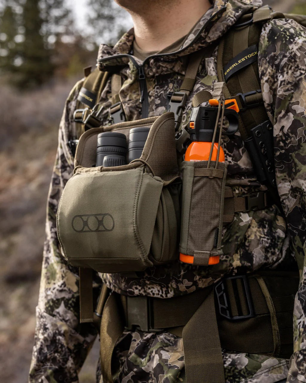 Eine Person in Camouflage trägt eine EBERLESTOCK Recon Bear Spray Pouch, hält ein Fernglas und eine orangefarbene Sprühdose in der Hand und steht im Freien, mit Bäumen und Hügeln im Hintergrund.