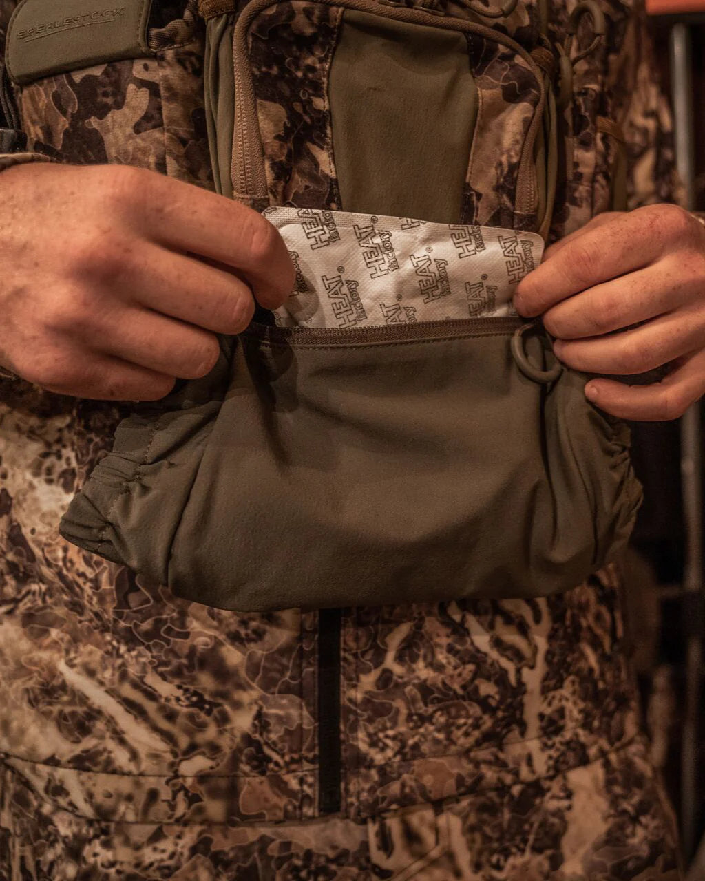 Ein Jäger in Camouflage legt einen EBERLESTOCK Recon Handwarmer in die Brusttasche eines Bino-Gurtes, damit die Hände warm und einsatzbereit bleiben.