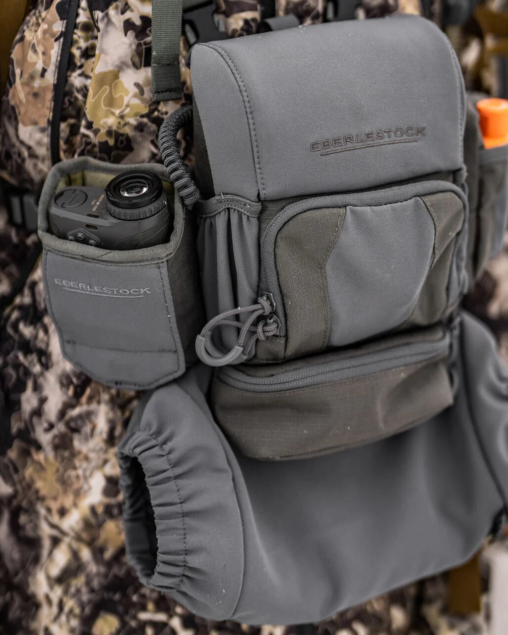 Nahaufnahme einer EBERLESTOCK Recon Rangefinder Pouch, die an einem Tarnrucksack befestigt ist, mit einer Kamera, die aus einer Tasche herausschaut, und orangefarbener Ausrüstung an der Seite.