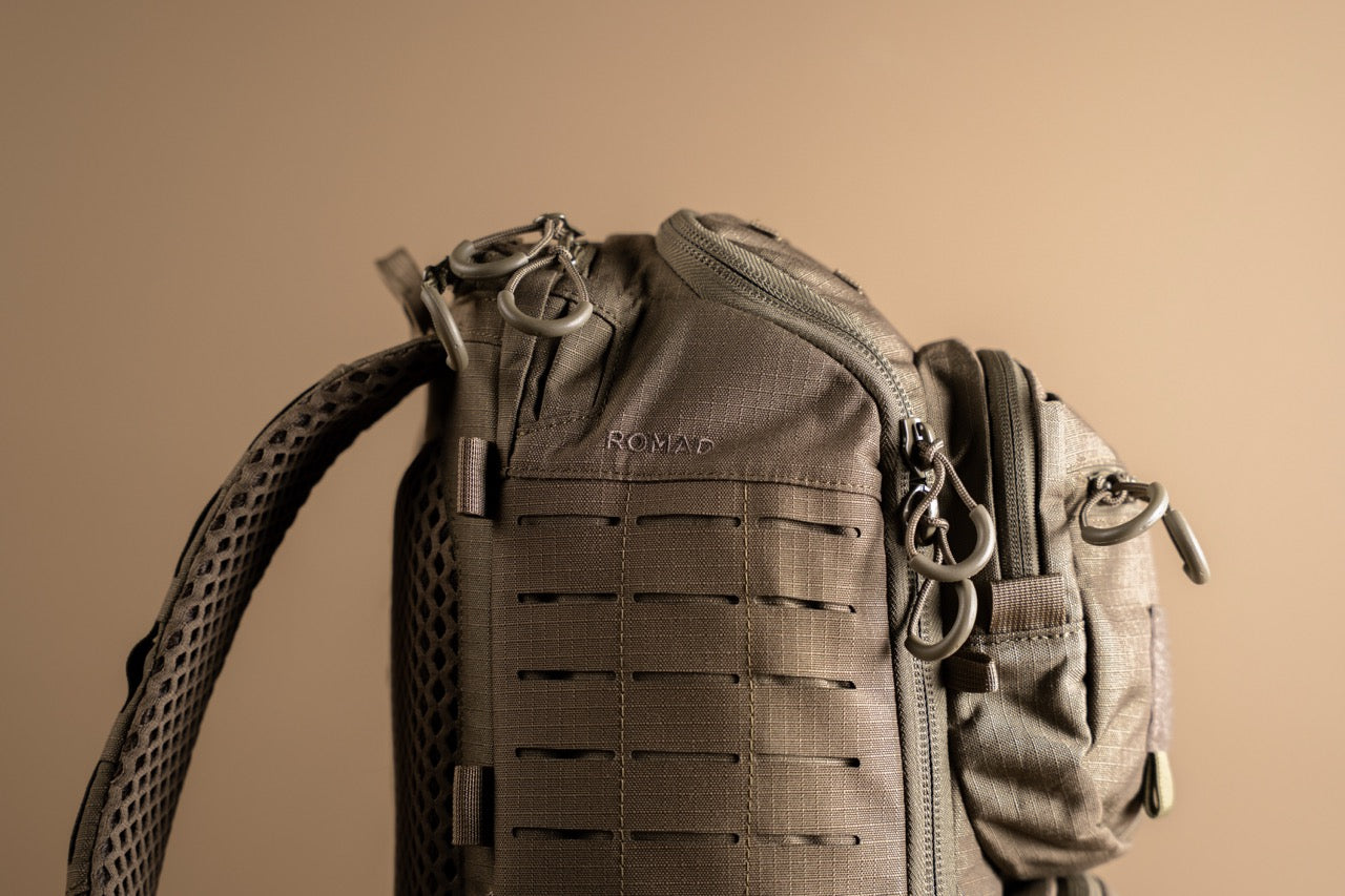Nahaufnahme eines schwarzen Eberlestock Romad Rucksacks von EBERLESTOCK, mit mehreren Reißverschlüssen, Schlaufen und MOLLE-Fächern. Der Schriftzug "ROMAD" ist auf den Stoff gestickt, der sich von einem einfarbigen Hintergrund abhebt.
