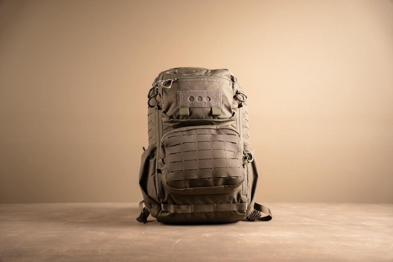 Ein robuster schwarzer EBERLESTOCK Romad Rucksack mit mehreren Fächern und MOLLE-Gurtband sitzt auf einer hellbraunen Oberfläche vor einem beigen Hintergrund.