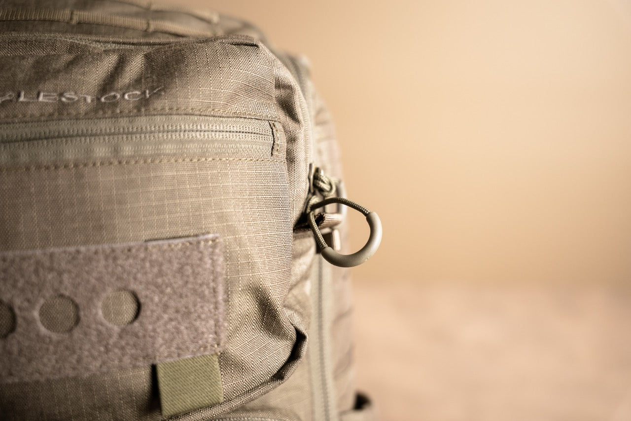 Nahaufnahme eines schwarzen EBERLESTOCK Eberlestock Romad taktischen Rucksacks, bei dem ein Metallring am Reißverschluss, die Stoffstruktur und ein Klettverschlussaufnäher auf der Vordertasche vor einem unscharfen neutralen Hintergrund hervorgehoben werden.