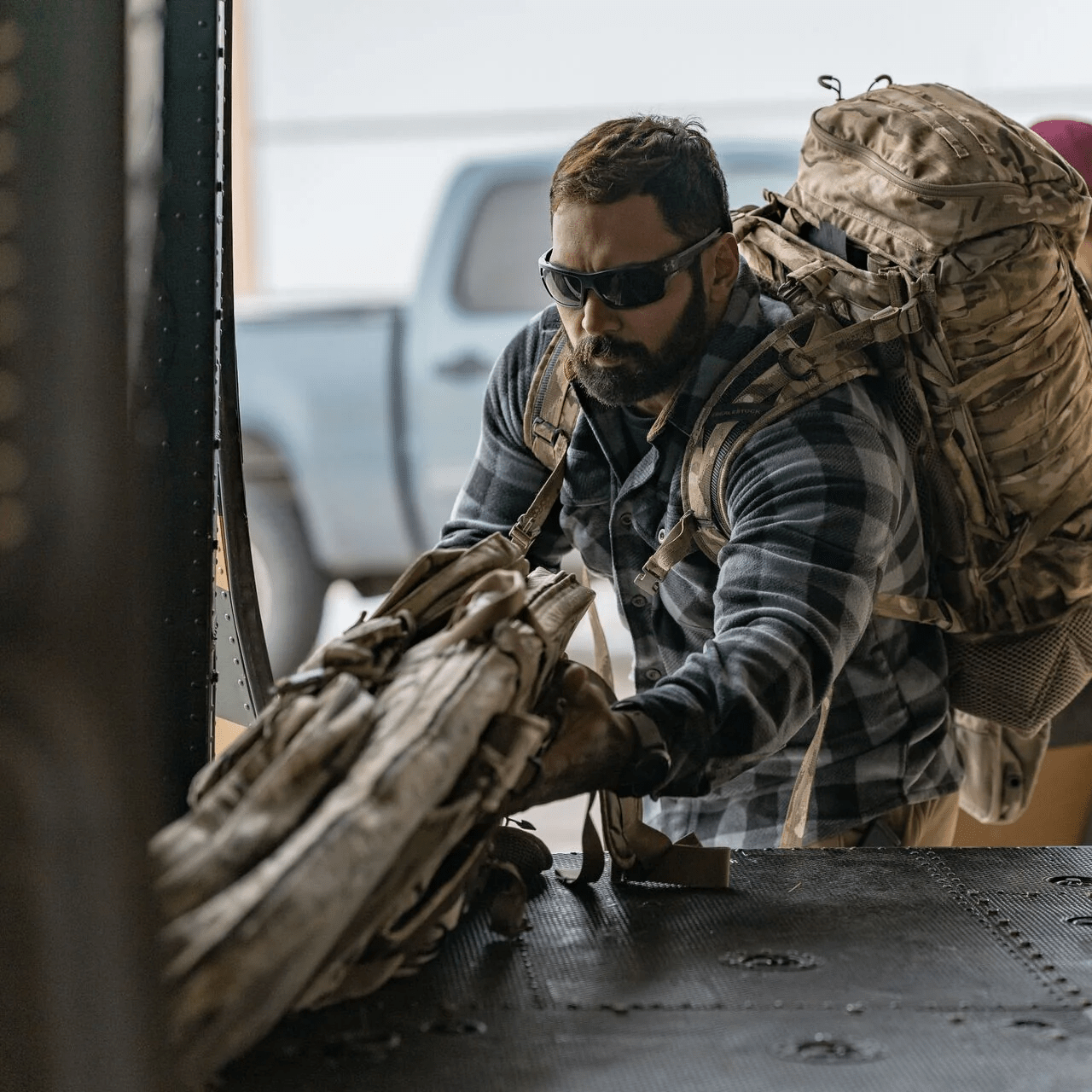 Ein Mann mit Sonnenbrille, kariertem Hemd und großem Rucksack lädt seine Ausrüstung - darunter die EBERLESTOCK Sniper Sled Drag Bag 52" - auf ein Fahrzeug oder Flugzeug, während im Hintergrund ein blauer Lastwagen zu sehen ist.