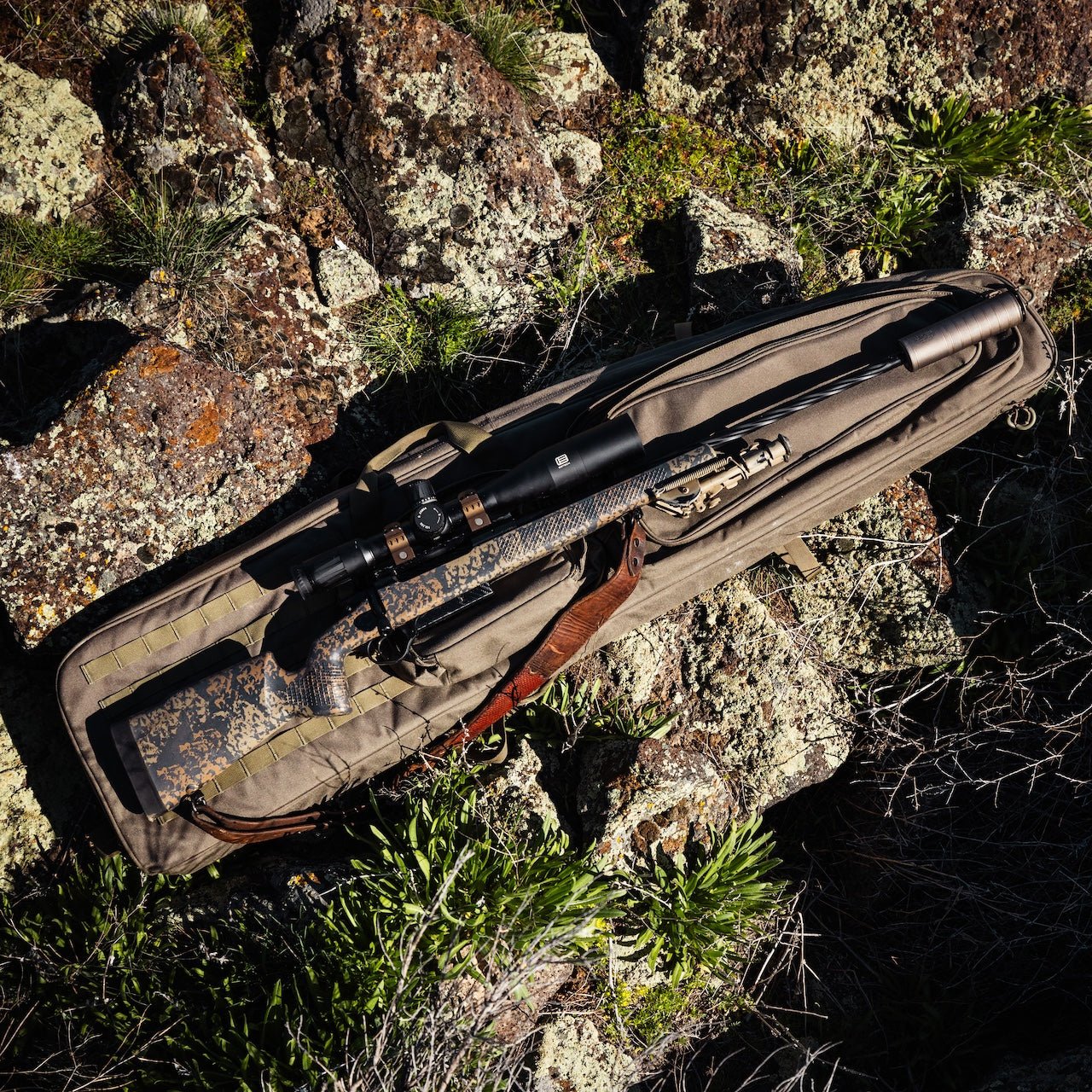 Ein Gewehr mit Zielfernrohr und Tarnmuster ruht in einer gepolsterten EBERLESTOCK Sniper Sled Drag Bag 57" auf felsigem Boden, umgeben von grünen Gras- und Moosflecken.